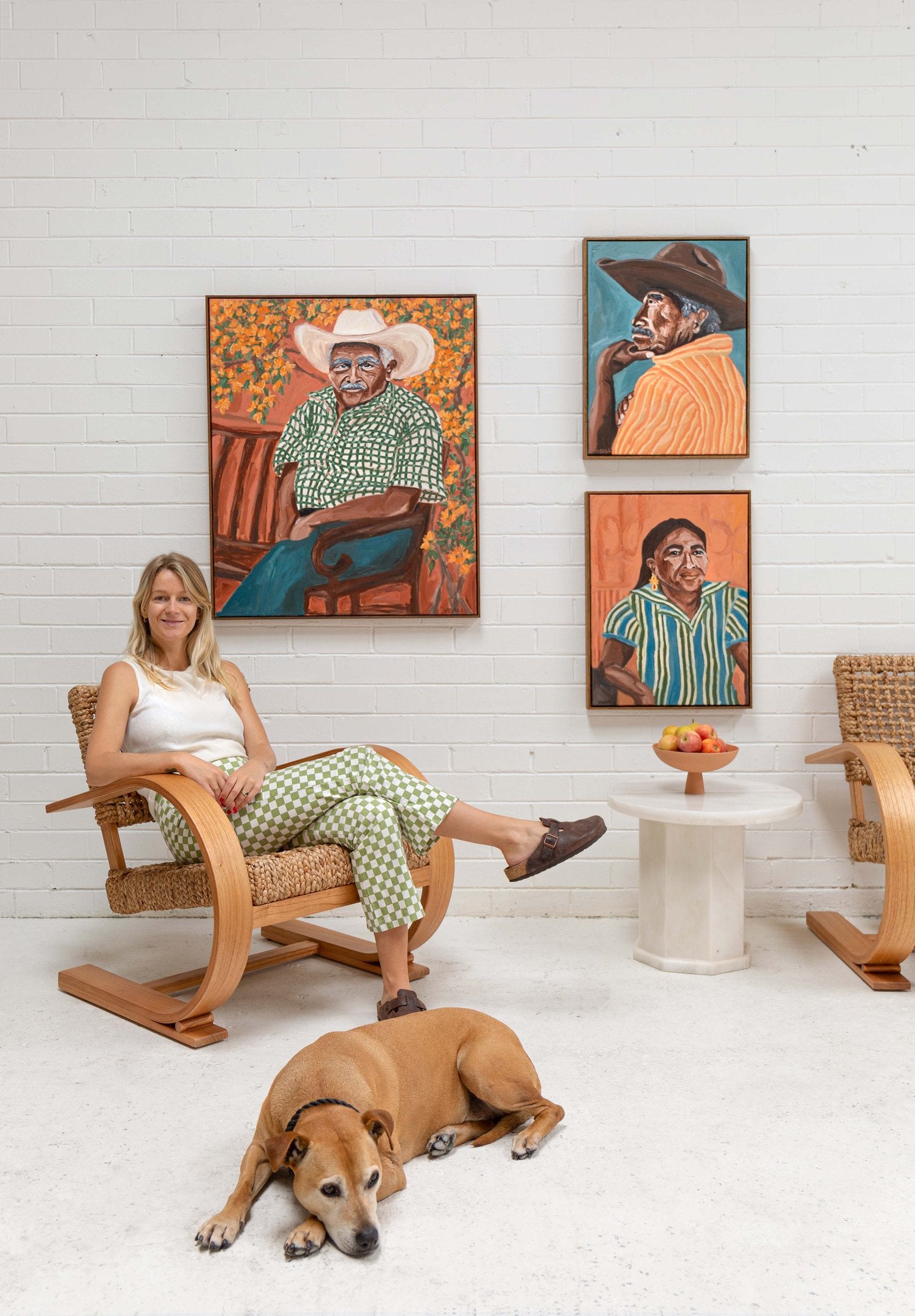 Artist Sammy Veall sitting in her studio with her dog with 3 paintings on the wall